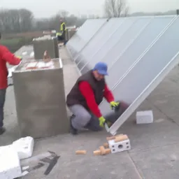 Montaż paneli słonecznych na dachu budynku; pracownicy w odzieży roboczej ustawiają panele, widoczne bloczki betonowe i cegły