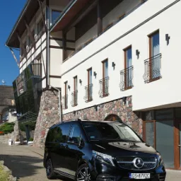 Czarny van Mercedes-Benz V-klasy zaparkowany przed budynkiem z kamienną elewacją i napisem 'Konferencje' na szklanych drzwiach.