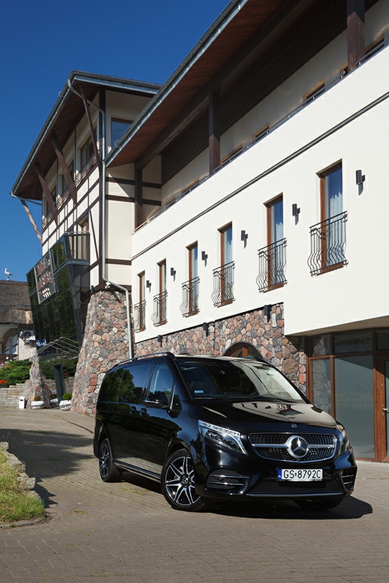 Czarny van Mercedes-Benz V-klasy zaparkowany przed budynkiem z kamienną elewacją i napisem 'Konferencje' na szklanych drzwiach.