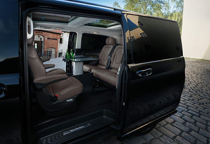 Wnętrze luksusowego, czarnego vana Mercedes-Benz z brązowymi skórzanymi siedzeniami, rozkładanym stolikiem z zielonymi butelkami i panoramicznym dachem, widok z otwartych przesuwnych drzwi...