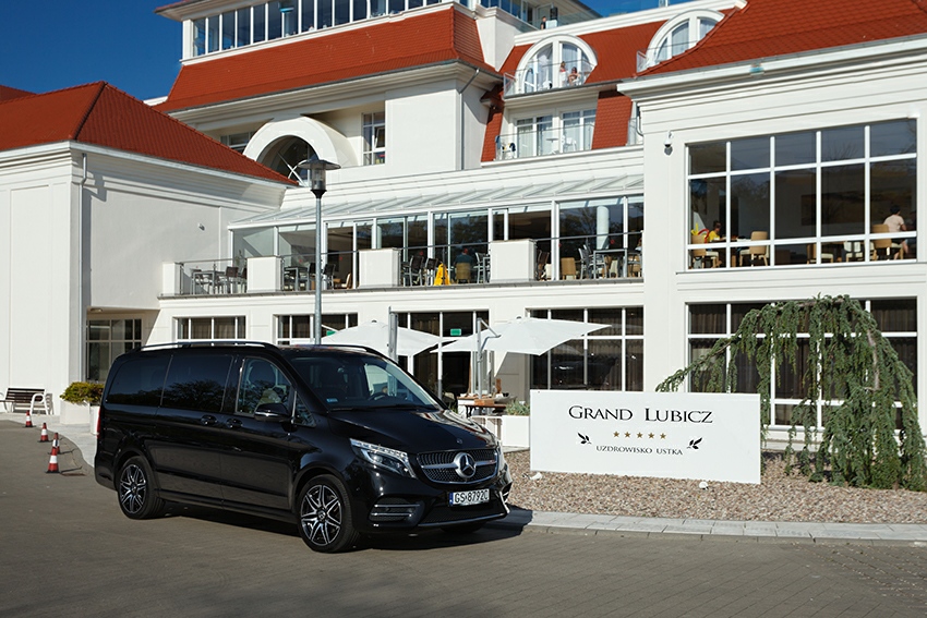 Czarny Mercedes-Benz V-klasy parkuje przed hotelem Grand Lubicz Uzdrowisko Ustka, widoczne stoły na balkonie i czerwone stożki drogowe.