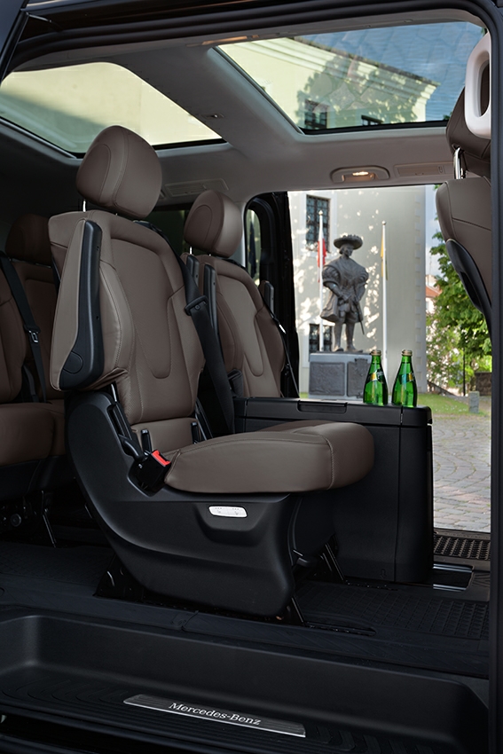 Wnętrze luksusowego vana z brązowymi skórzanymi siedzeniami, szklanym dachem i widokiem na pomnik na zewnątrz, dwie zielone butelki na stoliku.