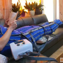 Osoba leżąca na kanapie, czytająca książkę podczas kompresyjnego masażu limfatycznego nóg za pomocą urządzenia z regulacją ciśnienia i czasu.