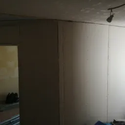 Montaż płyt karton gips na ścianie w trakcie remontu, widoczne profile i narzędzia w tle, surowy stan wykończenia.