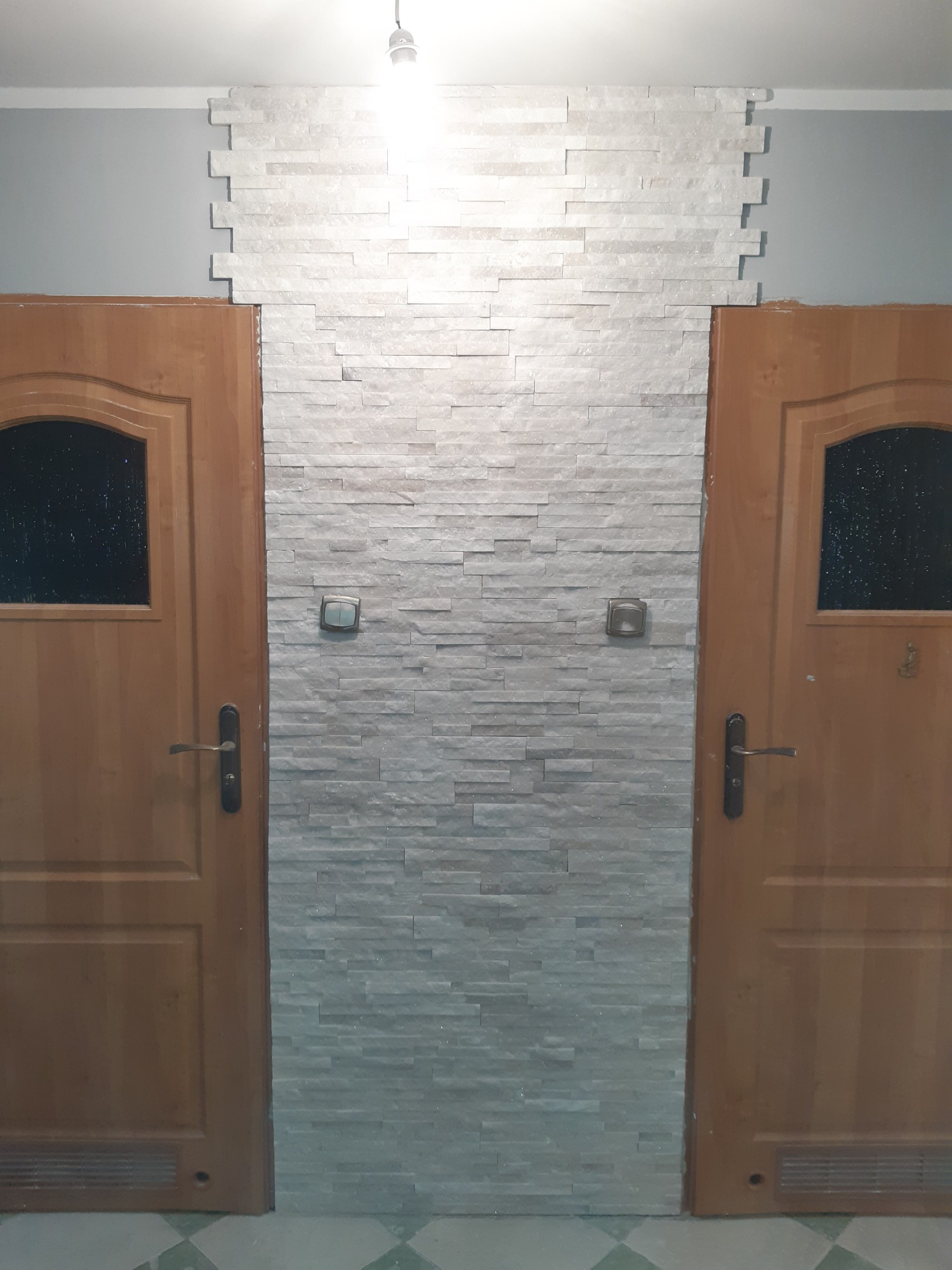 Ściana wykończona dekoracyjną płytką imitującą kamień, pomiędzy dwoma drewnianymi drzwiami z szybkami, widoczne włączniki światła.