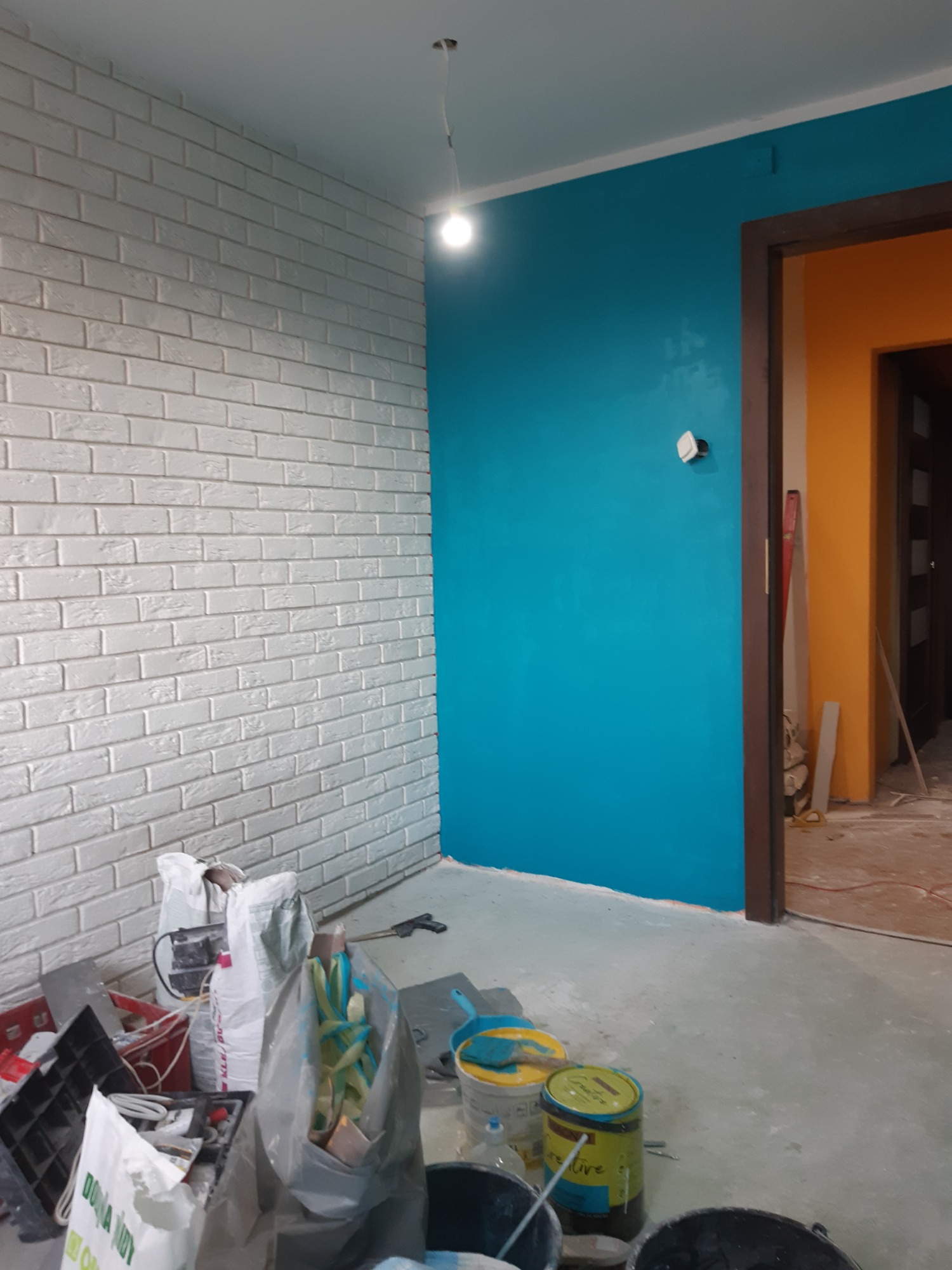 Wnętrze w trakcie remontu: ściana z białej cegły dekoracyjnej, błękitna ściana pomalowana farbą, widoczne puszki z farbą, worki z materiałami budowlanymi i narzędzia.