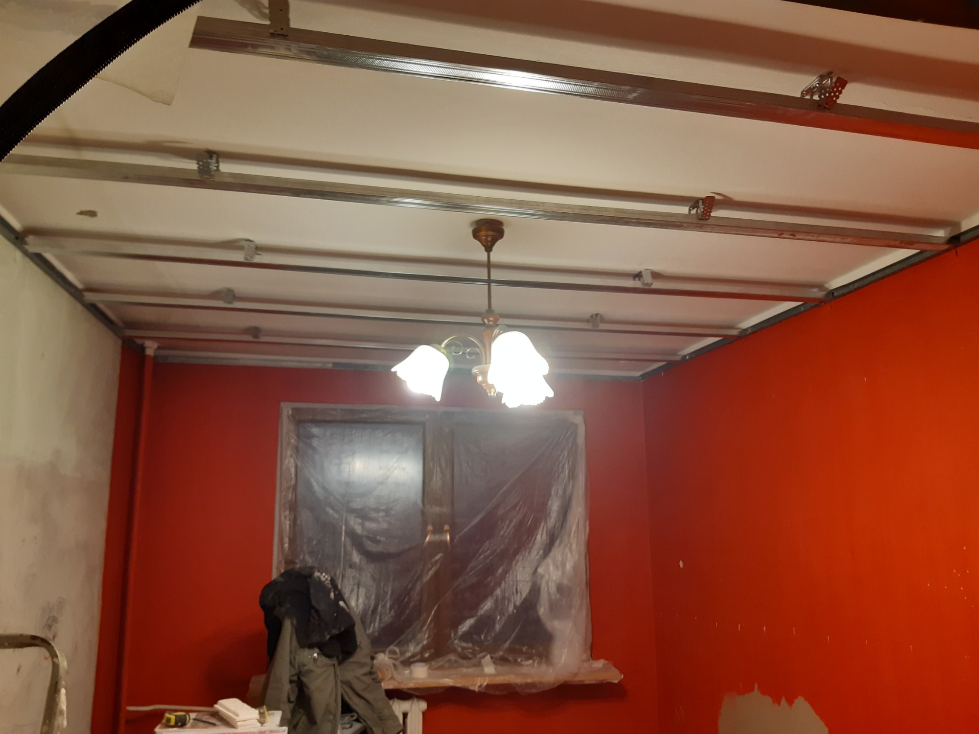 Montaż profili sufitowych podwieszanych w pokoju z pomalowanymi na czerwono ścianami, widoczny stary żyrandol i okno zabezpieczone folią malarską.