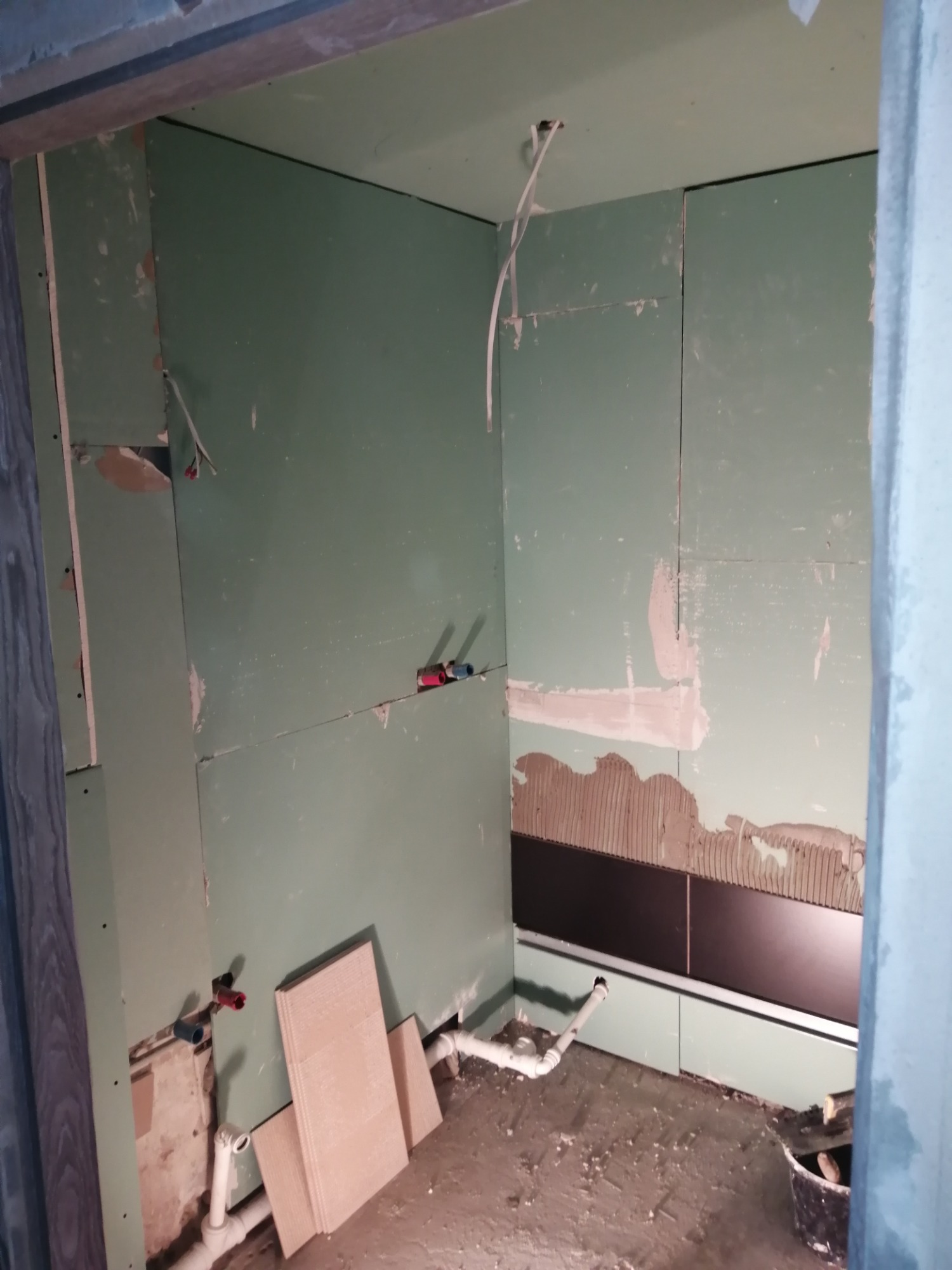 Wnętrze łazienki w trakcie remontu: widoczne płyty kartonowo-gipsowe na ścianach, częściowo położone ciemne płytki, wystające rury instalacji wodnej i przygotowane otwory pod instalację elektryczną.