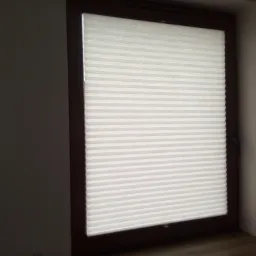 Zamknięta, biała plisa okienna w brązowej ramie okiennej, widoczny fragment ściany w szarym odcieniu i fragment podłogi.