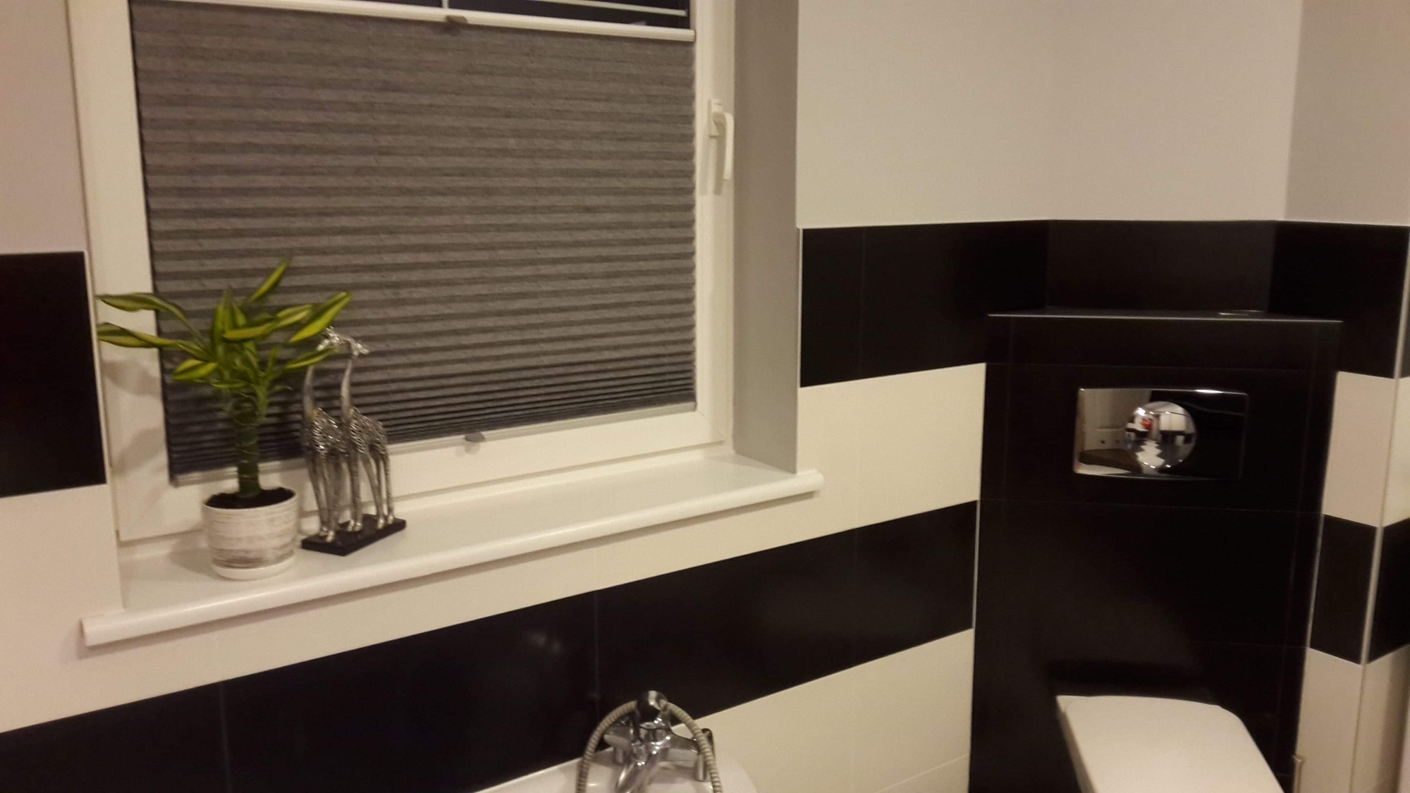 Szara plisa okienna zamontowana w oknie w łazience z czarno-białymi pasami na ścianach, na parapecie roślina w doniczce i dekoracja w kształcie żyraf.