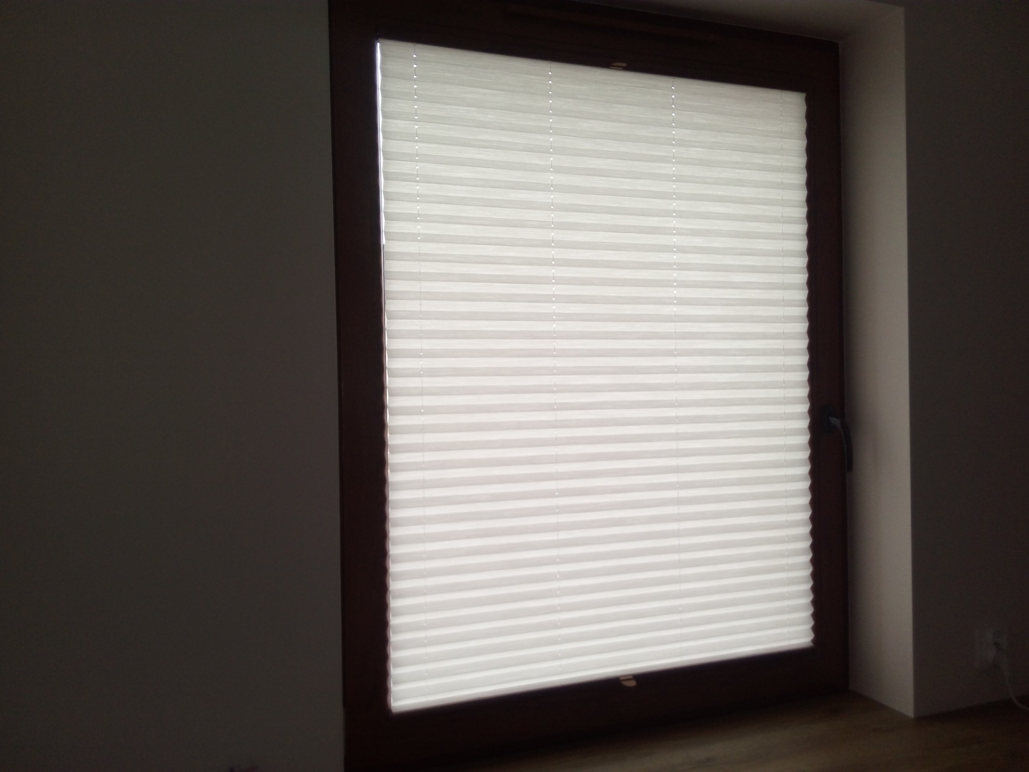 Zamknięta, biała plisa okienna w brązowej ramie okiennej, widoczny fragment ściany w szarym odcieniu i fragment podłogi.