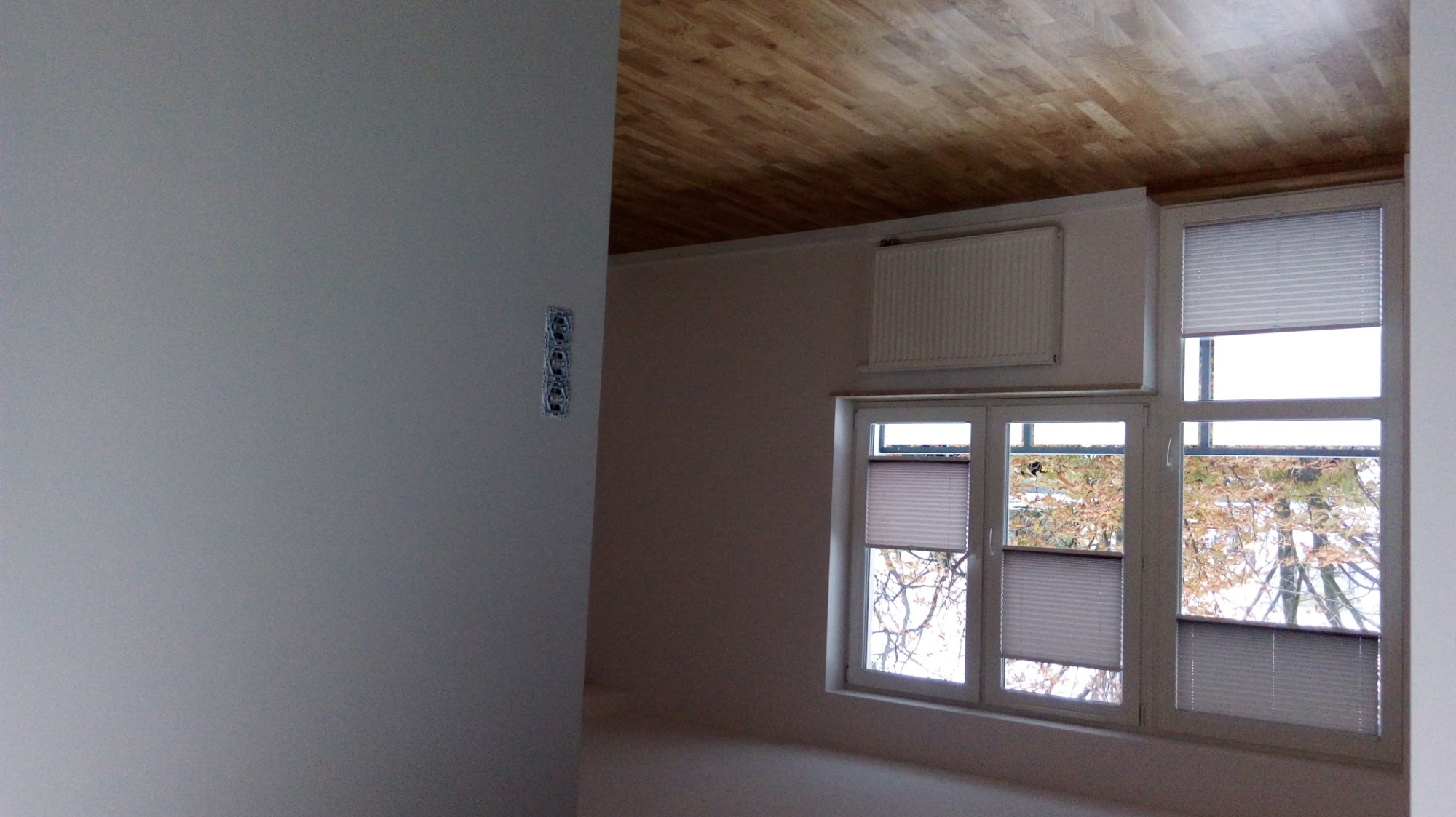 Wnętrze pokoju z zamontowanymi plisami okiennymi w trzech oknach, widok na zewnątrz z widocznymi drzewami, drewniany sufit i grzejnik pod oknem.