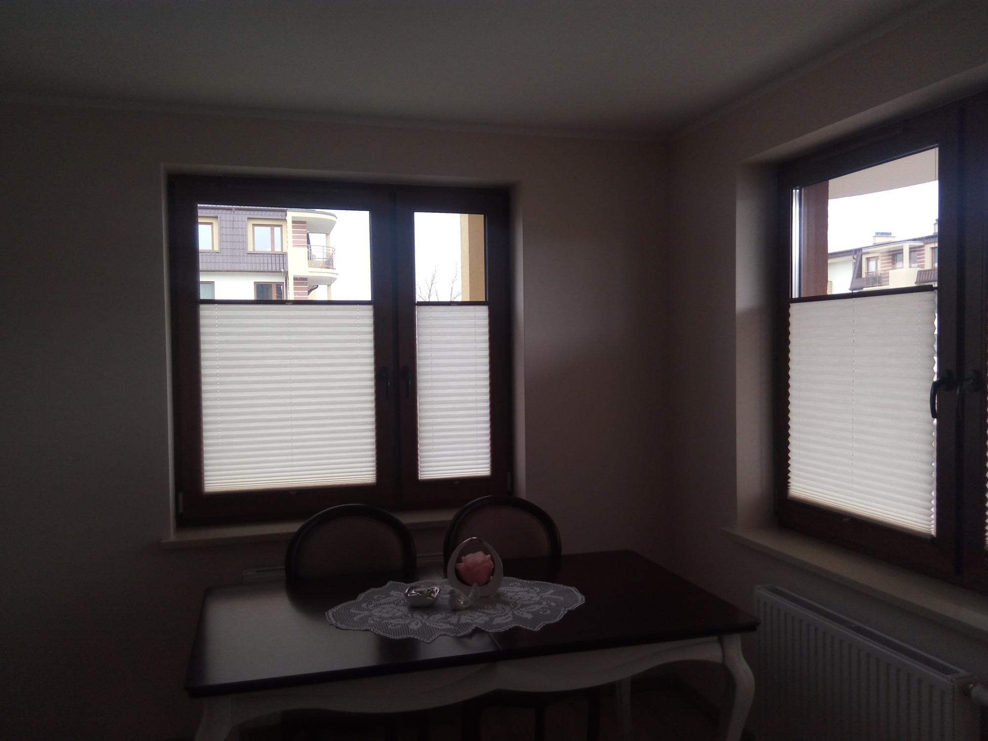 Dwa okna z plisowanymi roletami w brązowych ramach, widok na budynek z balkonami, stół z dekoracyjną serwetą i ozdobami.
