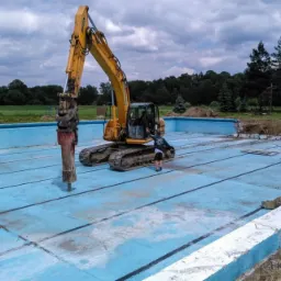 Żółta koparka z młotem pneumatycznym rozbija dno niebieskiego basenu, w tle drzewa i pochmurne niebo.