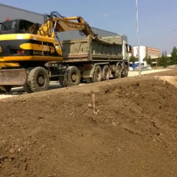 Żółto-czarna koparka na tle ciężarówki z muldą, w trakcie załadunku ziemi na placu budowy w słoneczny dzień.