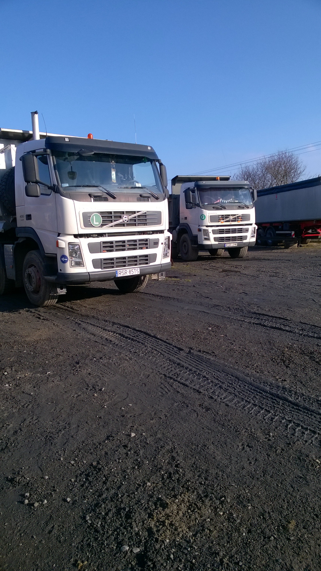 Dwa białe samochody ciężarowe Volvo z zielonym logo firmy na tle ciemnego, ubitego gruntu i błękitnego nieba.