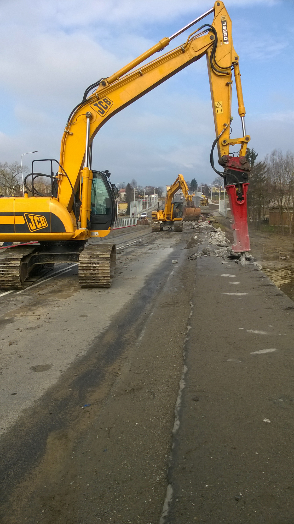 Żółty koparko-ładowarka JCB z czerwonym młotem hydraulicznym kruszy beton na powierzchni, w tle widoczna kolejna maszyna budowlana i zabudowania miejskie.
