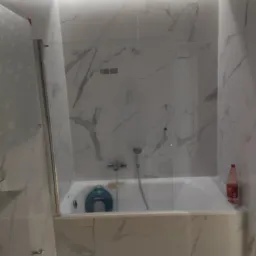 Wykończona łazienka z wanną obudowaną marmurowymi płytkami, szklany parawan nawannowy, niebieska deska sedesowa w wannie, butelka płynu do mycia na brzegu wanny.