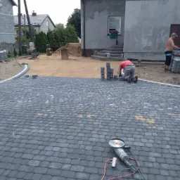 Prace brukarskie w toku: ułożona część nawierzchni z szarej kostki, pracownik na kolanach układa kolejne elementy, w tle przygotowany teren i sterta kostki, na pierwszym planie szlifierka kątowa.