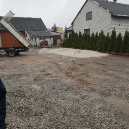 Przygotowanie terenu pod kostkę brukową: wywrotka z otwartą skrzynią, hałdy żwiru, częściowo utwardzony teren, szpadel oparty o rząd tuj, dom w tle, pochmurne niebo.