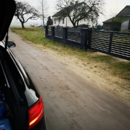 Nowoczesne, poziome ogrodzenie panelowe z betonowymi słupkami w kolorze grafitowym, widoczne z perspektywy otwartego bagażnika samochodu na polnej drodze, w tle dom i drzewa bez liści.