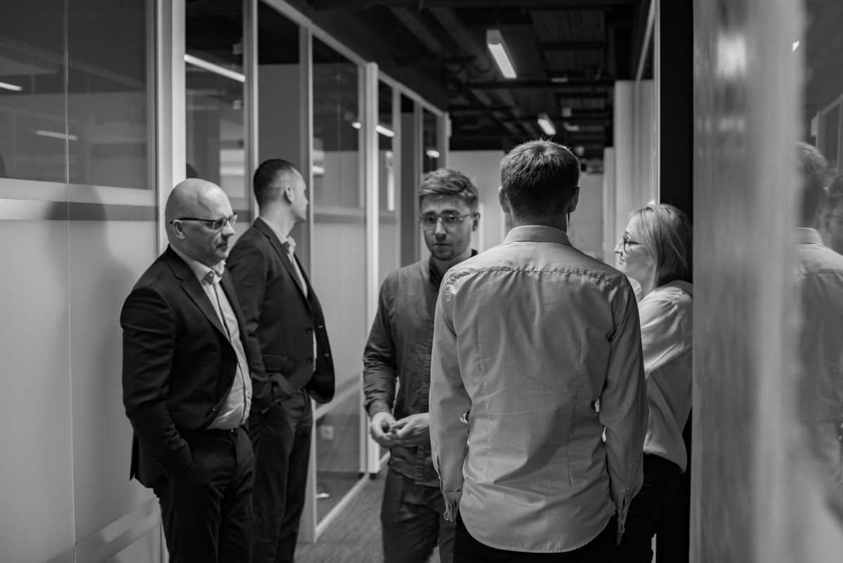 Czarno-białe zdjęcie przedstawia grupkę pięciu osób w biurowym korytarzu z przeszklonymi ścianami, gdzie jeden z mężczyzn ubrany w garnitur stoi z rękami w kieszeniach, a inny w szarej koszuli...