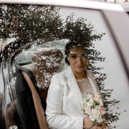 Karolina Krupiak Fotografia - Uśmiechnięta panna młoda w białym żakiecie, trzyma bukiet różowych i białych róż, siedząc w luksusowym samochodzie z brązową skórzaną tapicerką, z odbiciem drzewa na szybie.