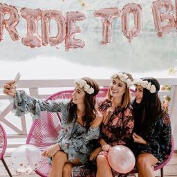 Karolina Krupiak Fotografia - Trzy uśmiechnięte kobiety w kwiatowych wiankach robią selfie na wieczorze panieńskim, siedząc na różowych krzesłach z balonami, na tle jeziora i napisu 'BRIDE TO BE' z balonów.