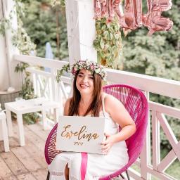 Karolina Krupiak Fotografia - Uśmiechnięta kobieta w białej sukience i wianku na głowie siedzi na różowym krześle na werandzie, trzymając białe pudełko z napisem 'Ewelina' w kolorze złotym; w tle balony foliowe z napisem 'BRIDE'.