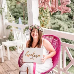 Uśmiechnięta kobieta w białej sukience i wianku na głowie siedzi na różowym krześle na werandzie, trzymając białe pudełko z napisem 'Ewelina' w kolorze złotym; w tle balony foliowe z napisem 'BRIDE'.