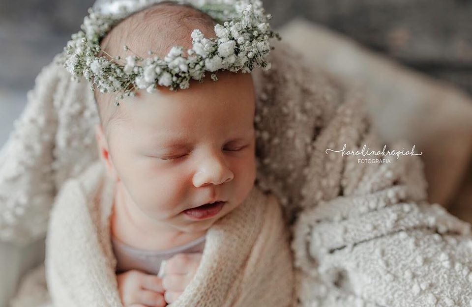 Śpiący noworodek w białym otulaczu i wianku z gipsówki na głowie, ułożony na puszystym, teksturowanym kocyku, z widocznym logo fotografa w prawym rogu.