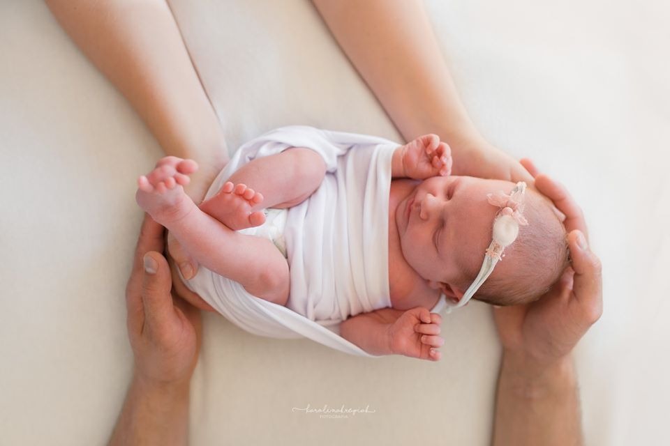 Noworodek owinięty w białą tkaninę, trzymany w dłoniach rodziców, z delikatną opaską na głowie z małym kwiatkiem, na jasnym tle.