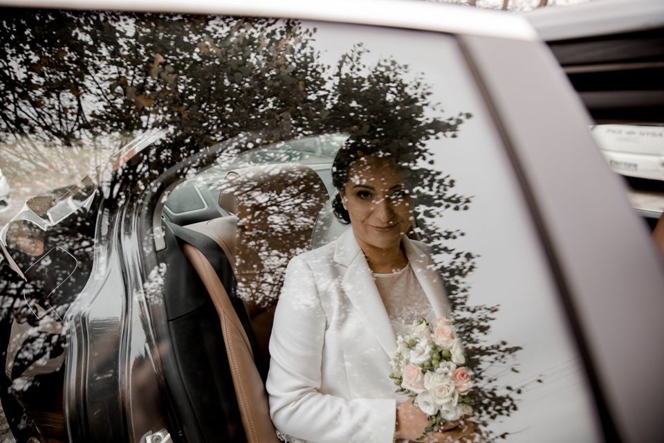 Uśmiechnięta panna młoda w białym żakiecie, trzyma bukiet różowych i białych róż, siedząc w luksusowym samochodzie z brązową skórzaną tapicerką, z odbiciem drzewa na szybie.