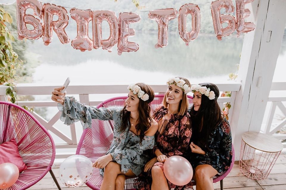 Trzy uśmiechnięte kobiety w kwiatowych wiankach robią selfie na wieczorze panieńskim, siedząc na różowych krzesłach z balonami, na tle jeziora i napisu 'BRIDE TO BE' z balonów.