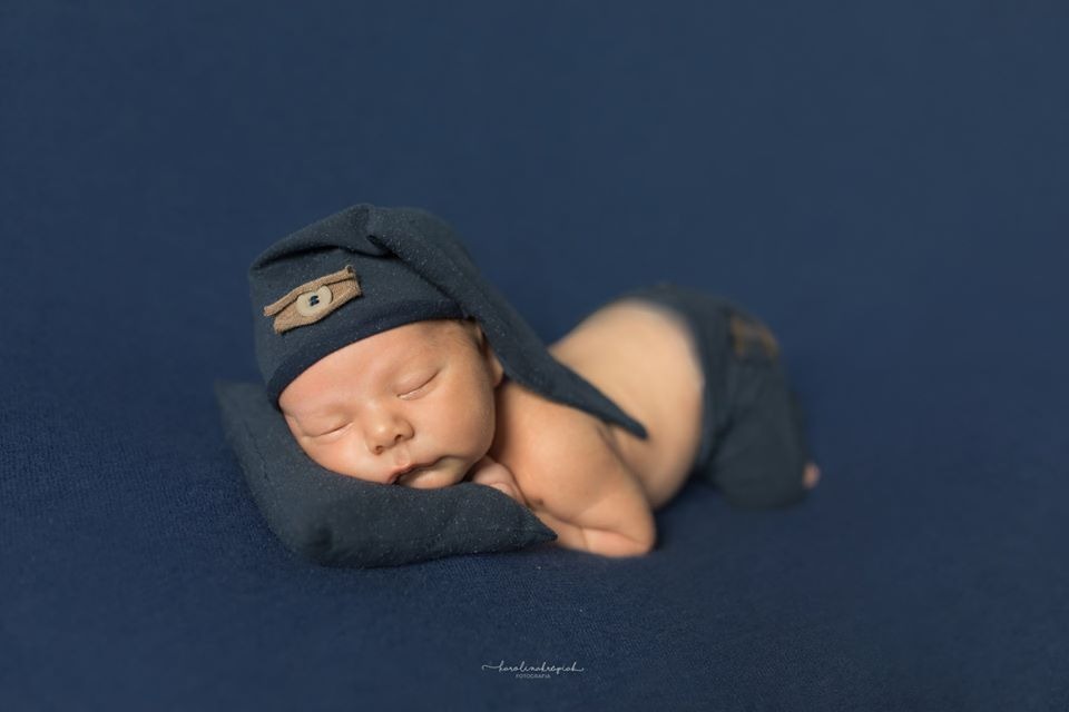 Śpiący noworodek w ciemnoniebieskim stroju z czapeczką z guzikiem, leżący na małej poduszce na jednolitym tle.