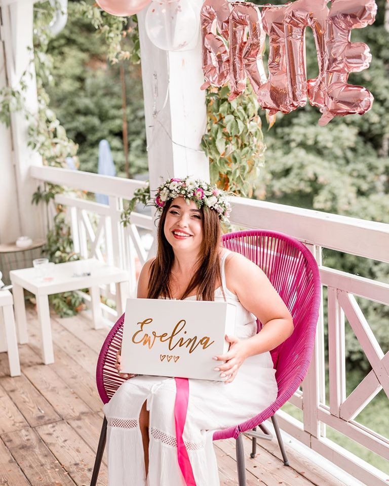 Uśmiechnięta kobieta w białej sukience i wianku na głowie siedzi na różowym krześle na werandzie, trzymając białe pudełko z napisem 'Ewelina' w kolorze złotym; w tle balony foliowe z napisem 'BRIDE'.