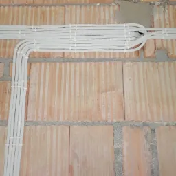 Białe przewody elektryczne ułożone w bruzdach ceglanej ściany, zabezpieczone plastikowymi opaskami, widoczne napisy na przewodach.