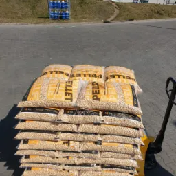 Stos worków z pelletem ułożonych na palecie, widoczny wózek paletowy, w tle budynki przemysłowe i rząd niebieskich butli gazowych.