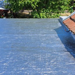 AT hydroizolacje Sp. z o.o. (suchachata.pl) - Błyszcząca, nowa izolacja bitumiczna na płaskim dachu, widok z góry, z fragmentem dachu z dachówki ceramicznej po prawej stronie i fragmentem budynku w tle.