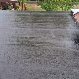 AT hydroizolacje Sp. z o.o. (suchachata.pl) - Płaska powierzchnia dachu pokryta czarną, błyszczącą gumą w płynie, widoczne połączenie z dachem krytym czerwoną dachówką ceramiczną, w tle zieleń drzew i budynki.