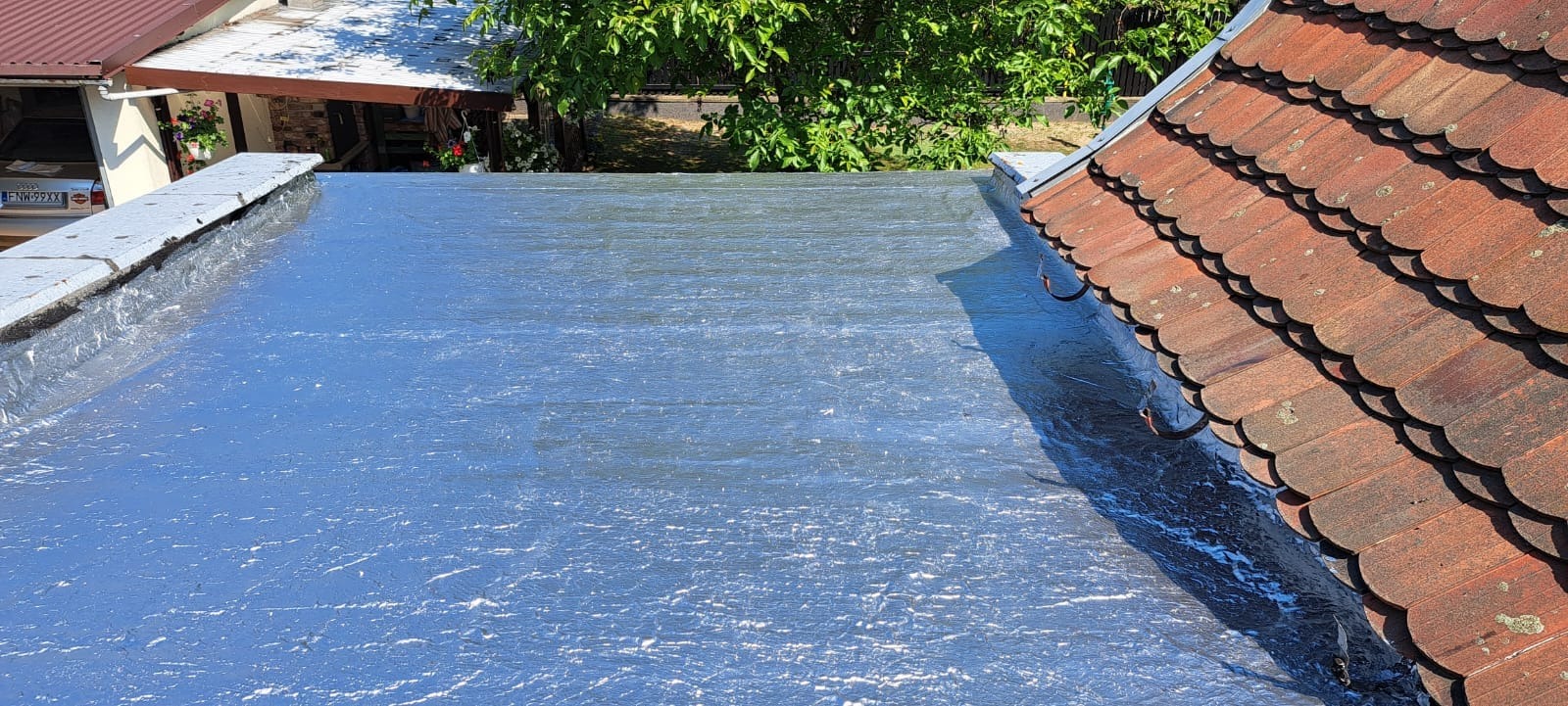 Błyszcząca, nowa izolacja bitumiczna na płaskim dachu, widok z góry, z fragmentem dachu z dachówki ceramicznej po prawej stronie i fragmentem budynku w tle.