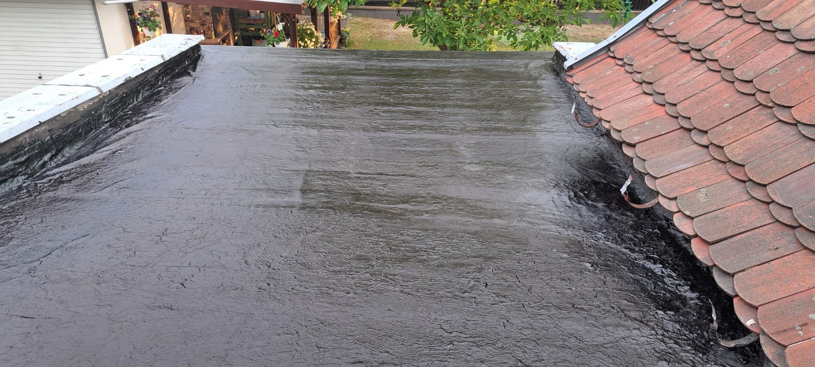 Płaska powierzchnia dachu pokryta czarną, błyszczącą gumą w płynie, widoczne połączenie z dachem krytym czerwoną dachówką ceramiczną, w tle zieleń drzew i budynki.