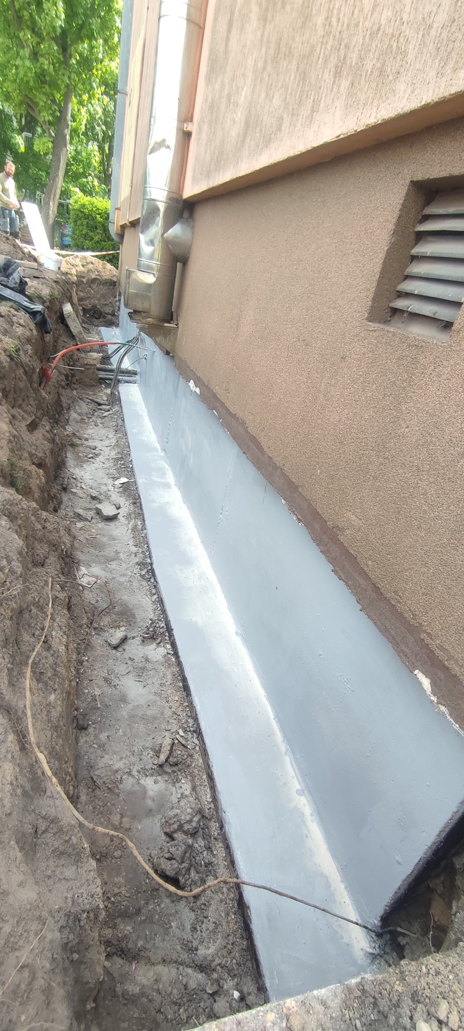 Szara hydroizolacja fundamentów, widoczna w wykopie przy ścianie budynku z brązowym tynkiem. Fragment instalacji i wentylacji. Praca w toku.