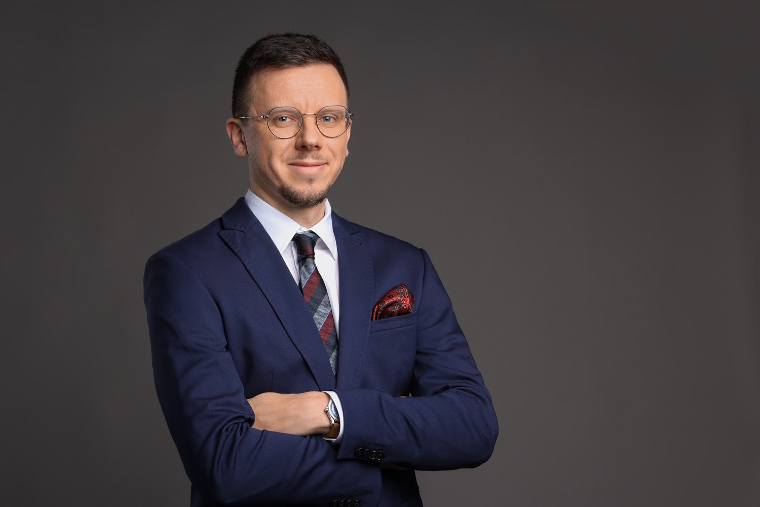 Portret uśmiechniętego mężczyzny w okularach, granatowym garniturze, białej koszuli i krawacie na szarym tle. Ręce skrzyżowane, widoczna poszetka w kieszonce.