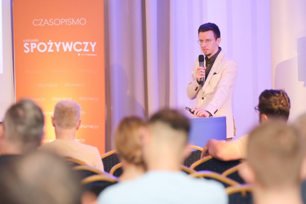 Prelegent w beżowym garniturze przemawia do publiczności podczas konferencji. W tle pomarańczowy baner z napisem 'Czasopismo Spożywczy'.