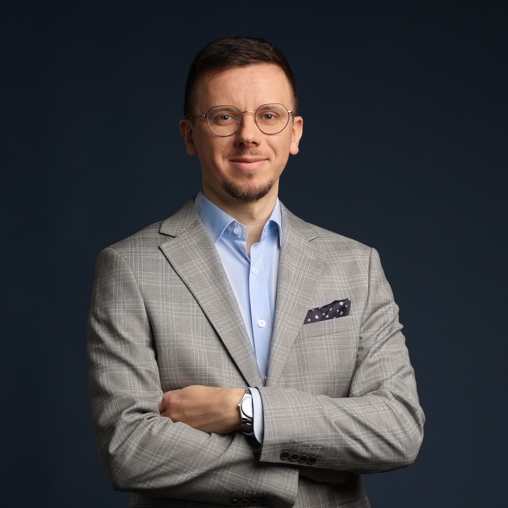 Portret mężczyzny w okularach i szarej marynarce w kratę, z poszetką w kieszonce, na ciemnoniebieskim tle. Skrzyżowane ręce, widoczny zegarek.