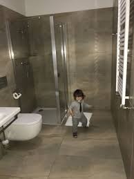 Chłopiec w szarym ubraniu z krawatem stoi w nowocześnie wykończonej łazience z kabiną prysznicową, umywalką z toaletą i grzejnikiem drabinkowym na tle szarych płytek.