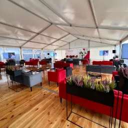 Wnętrze namiotu eventowego z drewnianą podłogą, czerwonymi i szarymi sofami, metalowymi kwietnikami z wrzosami, ekranami wyświetlającymi prezentacje i sceną w tle.