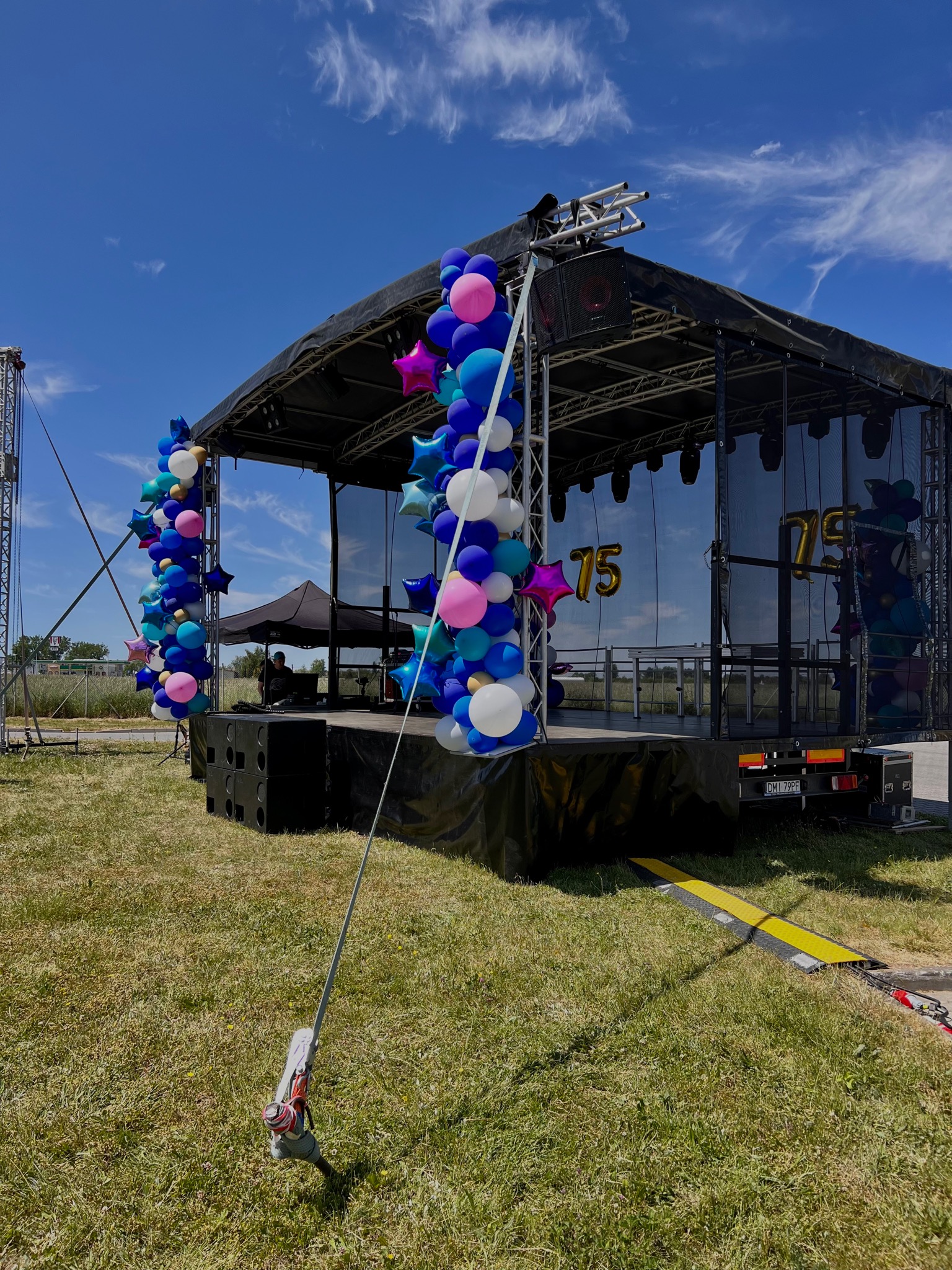 Scena plenerowa z nagłośnieniem, oświetleniem i dekoracją balonową w kolorach niebieskim, różowym i białym. W tle błękitne niebo z chmurami. Na scenie widoczne balony w kształcie cyfr '7' i '5'.