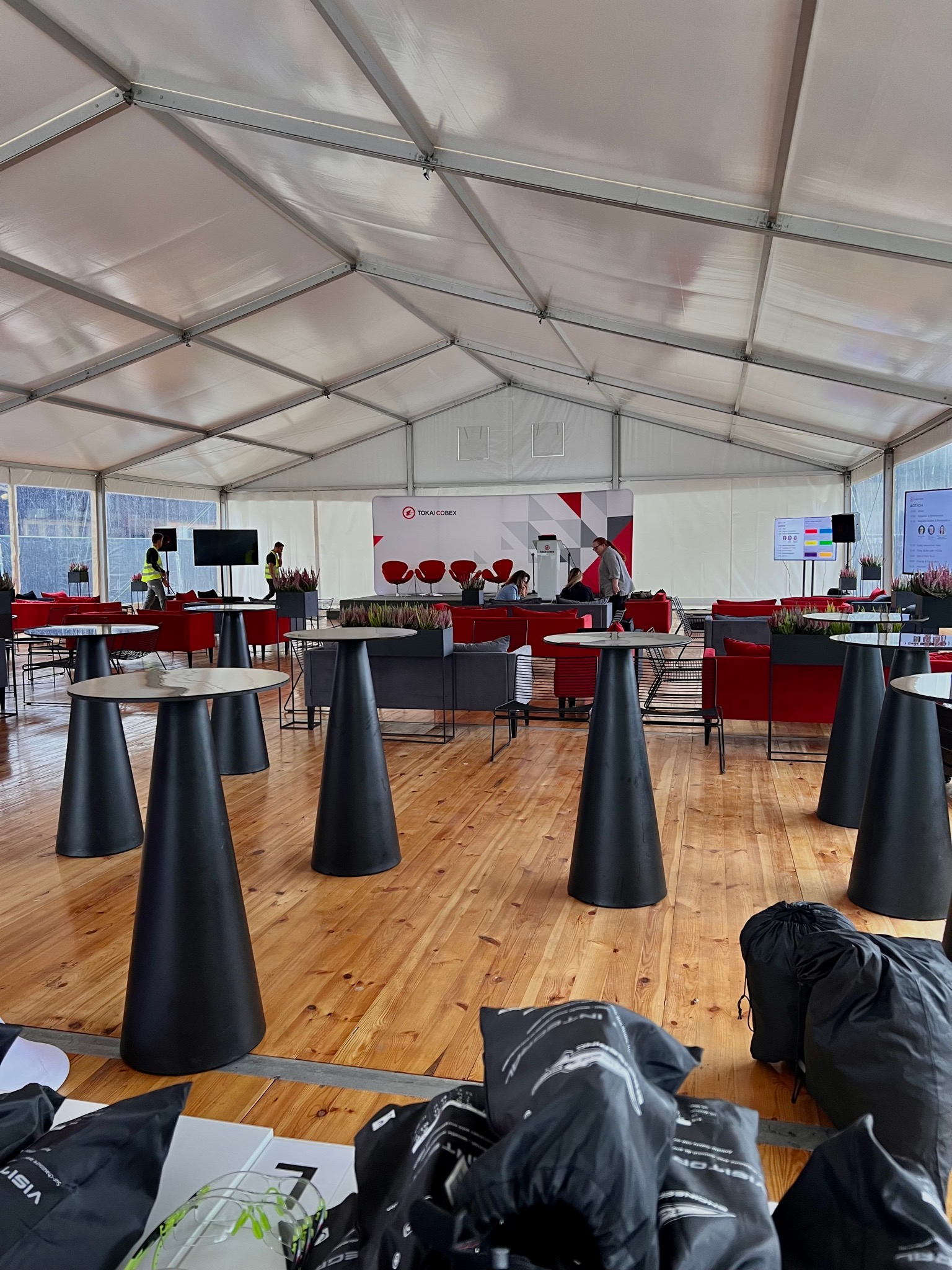 Wnętrze namiotu eventowego z drewnianą podłogą, czarnymi stolikami-stożkami, czerwonymi sofami i ekranami. W tle widoczny baner z logo firmy Tokai Cobex oraz osoby przygotowujące przestrzeń.
